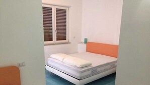 Une petite chambre propre, avec un lit simple, une fenêtre avec des stores, une table de chevet et une petite commode.