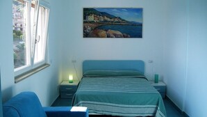 A bedroom with a bed, a chair, a nightstand, a bedside table, and a painting of a coastal scene. - Studio apartment Mirtillo (Pisciotta)
