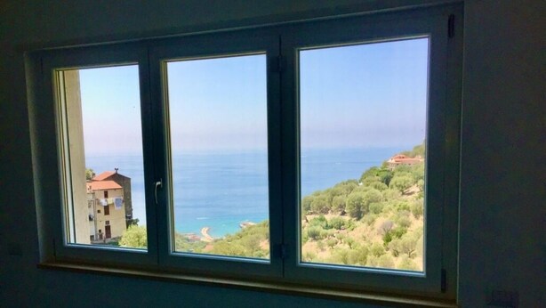 A room with a large window overlooking a coastal landscape, including a building and trees. - Studio apartment Mirtillo (Pisciotta)