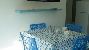 A dining area with a table covered in a blue and white patterned tablecloth, surrounded by blue chairs. A television is mounted on the wall above the table. - Studio apartment Mirtillo (Pisciotta)