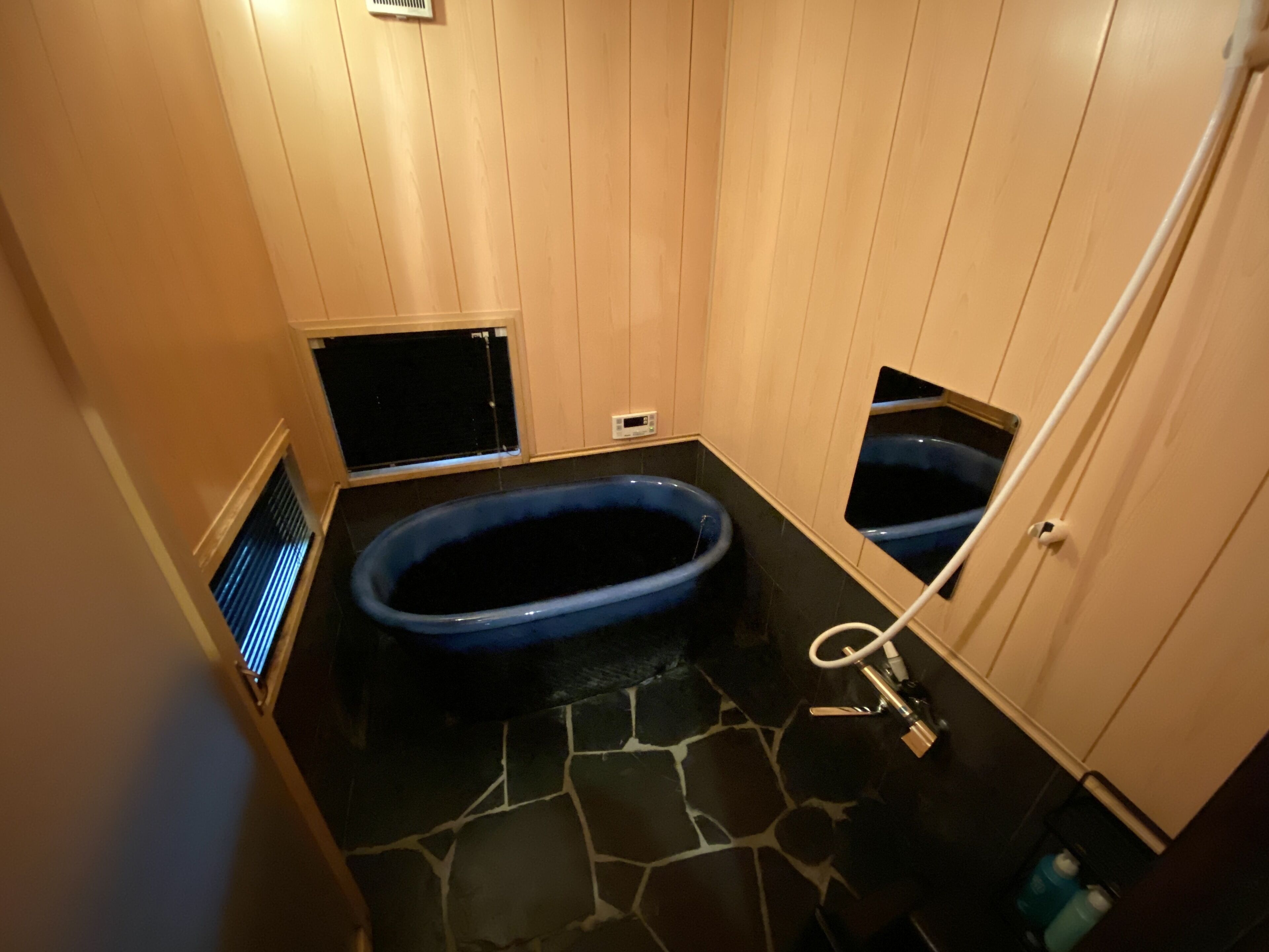 A small, dimly lit bathroom with a blue bathtub, a stone floor, and wooden walls.