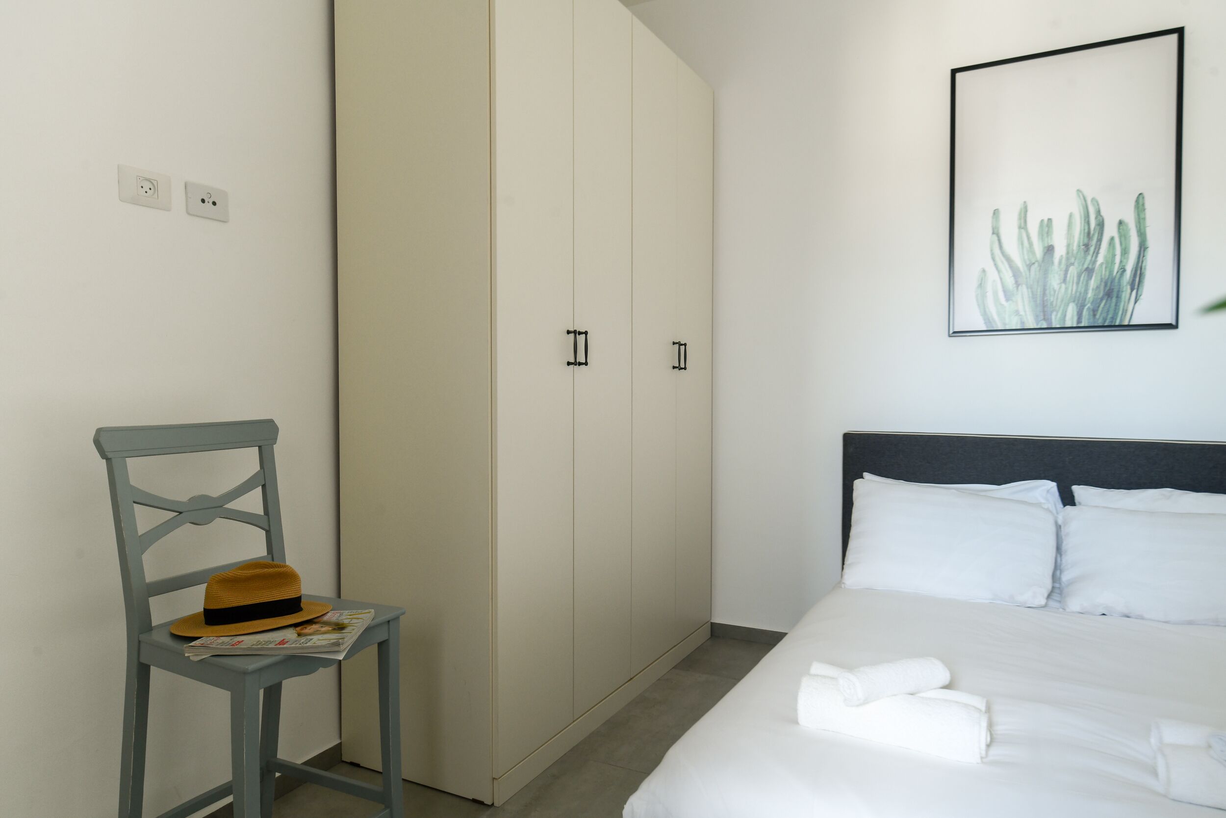 Un dormitorio con una cama, un armario, una silla con un sombrero y un cuadro en la pared.