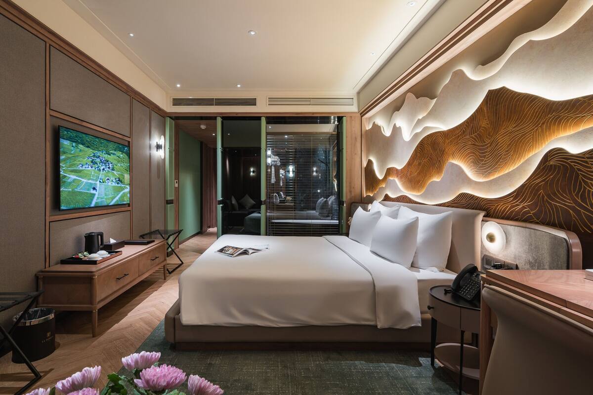 A modern hotel room with a large bed, a TV, a desk, and a unique wall design.