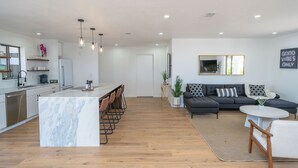 Eine moderne Küche mit einer Marmorinsel, ein Essbereich mit Stühlen, ein Wohnzimmer mit einer Couch und einem Couchtisch sowie ein Schild mit der Aufschrift 'GOOD VIBES ONLY'.