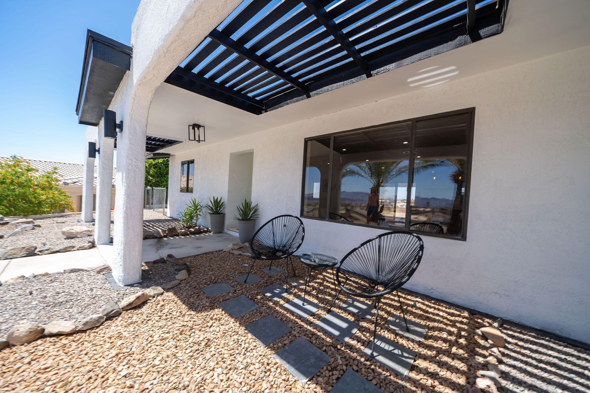 Een modern buitenterras met een pergola, twee zwarte draadstoelen en uitzicht op het woestijnlandschap door een groot raam.