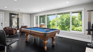 A modern living room with a pool table, leather chairs, and large windows offering a view of trees.