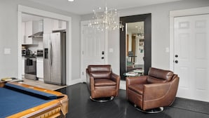 A modern living room with a pool table, two leather armchairs, a chandelier, and a kitchen in the background.