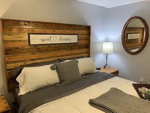 A bedroom with a wooden headboard, a bed with gray bedding, a bedside lamp, and a mirror on the wall.