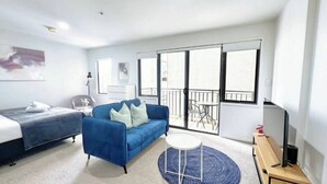 A modern hotel room with a bed, a blue sofa, a small round table, and a balcony with outdoor furniture. - Convenient Studio Steps from AUT University (Auckland)