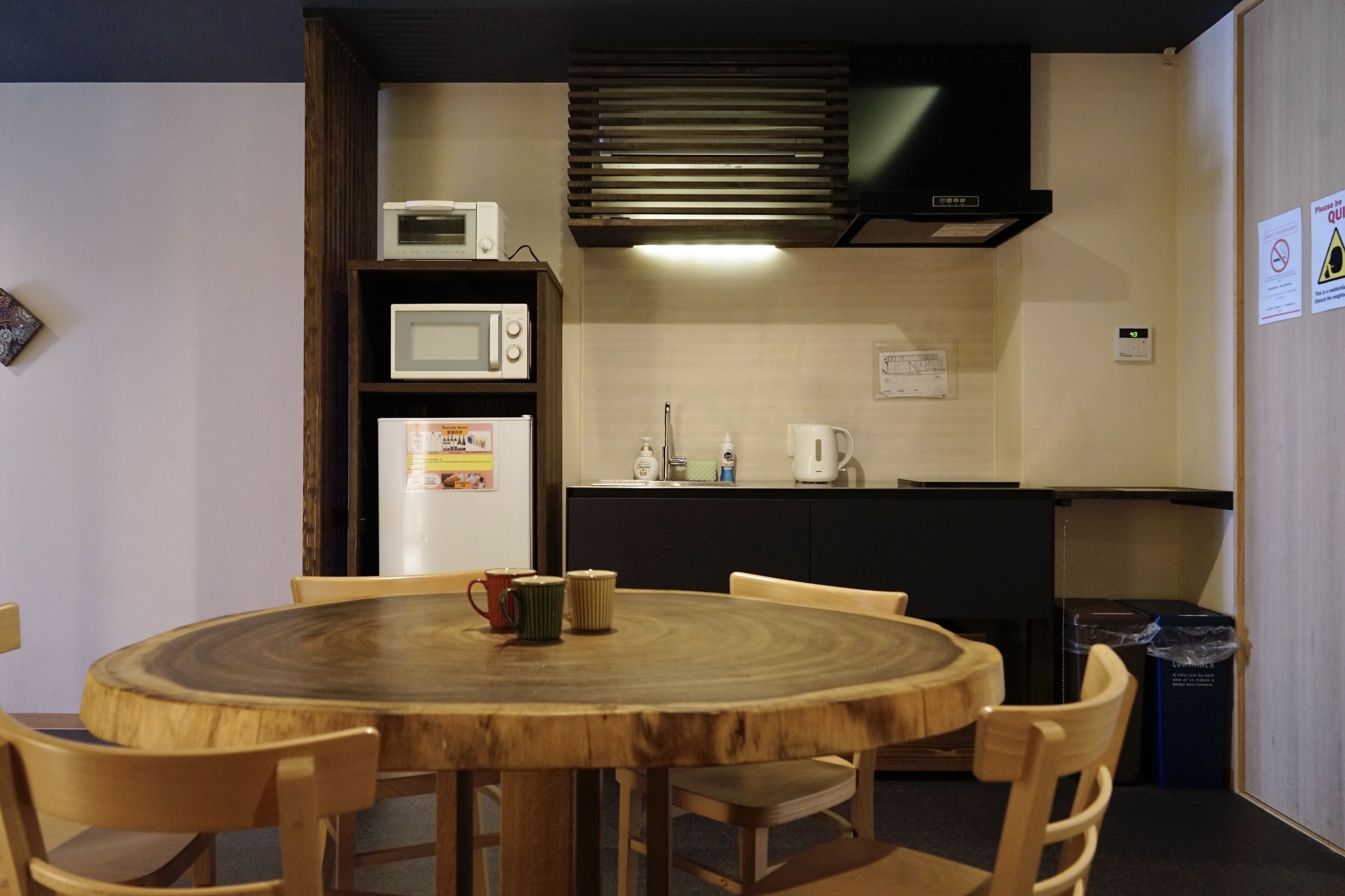 A compact kitchen with a microwave, refrigerator, and coffee maker. There is a round wooden table with chairs, and a trash bin is visible.