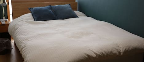 Una cama bien tendida con cabecera de madera, dos almohadas, una lámpara de noche y una mesita de noche con un teléfono y un bolso.