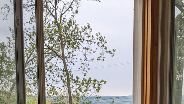 A corner room with a wicker chair and a view of a green landscape through large windows. - Casa Ceylon by Lake Seneca (Dundee)