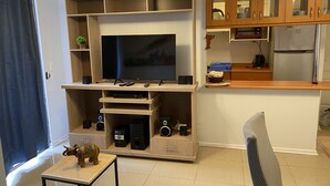 Een woonkamer met een flatscreen televisie, een houten dressoir, een keukenhoek met kastjes en een kleine tafel met een decoratief olifantje.