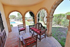 Un patio techado con mesa y sillas, arcos, y vistas a un jardín y una piscina.