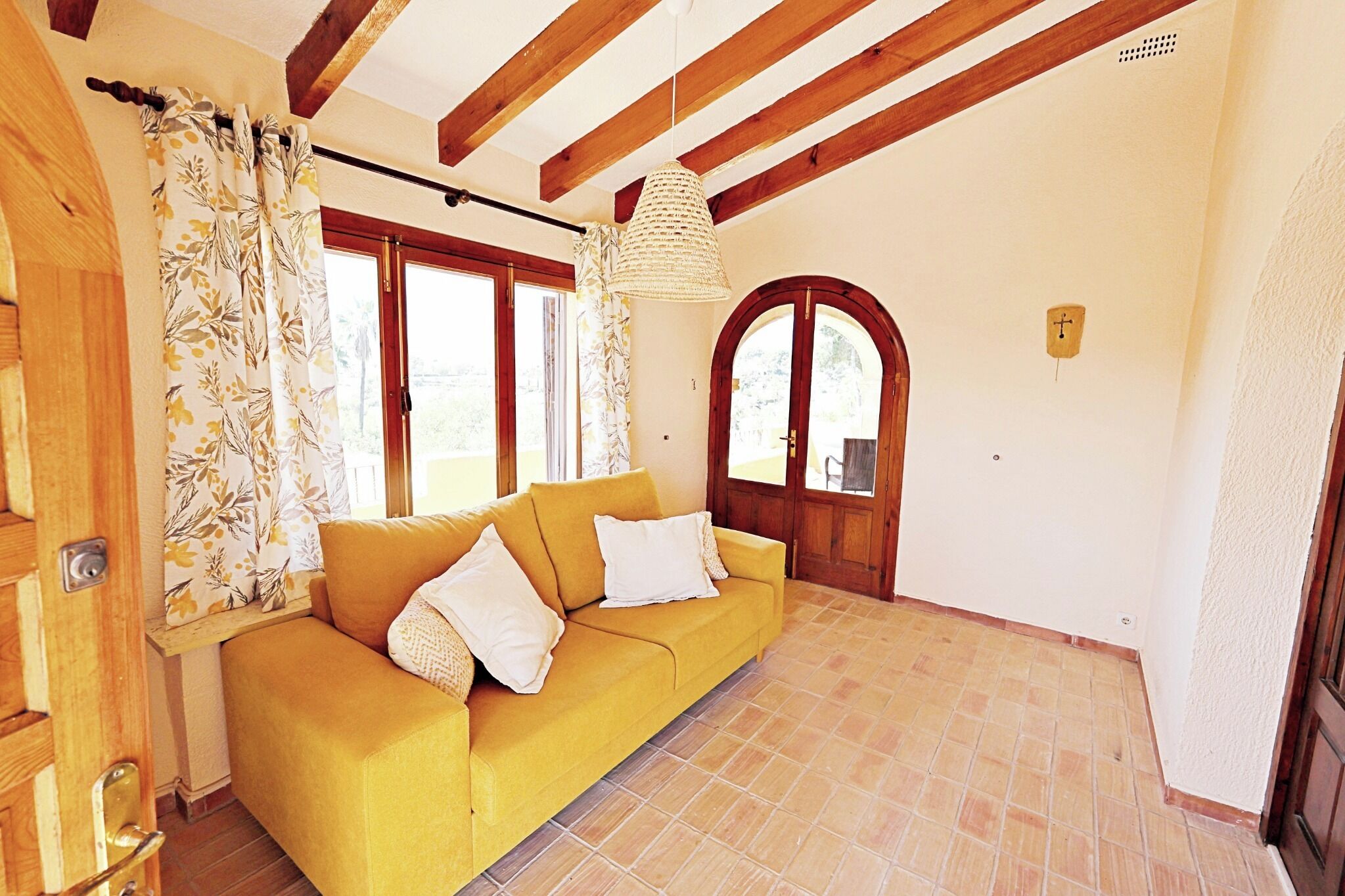 Un sofá amarillo con cojines blancos en una habitación con vigas de madera, una lámpara de fibra natural y una puerta con un ventanal de arco.