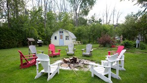 Un jardin avec un foyer entouré de fauteuils Adirondack, un petit abri de jardin et une maison en arrière-plan.