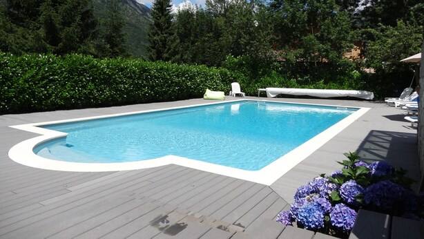 Une piscine rectangulaire bordée de blanc, entourée d’une terrasse en bois. On y trouve des chaises longues et un espace abrité avec une table.