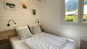 Une chambre à coucher avec une tête de lit en bois, deux lampes fixées au mur, un lit avec une literie blanche et une fenêtre donnant sur un paysage verdoyant et montagneux.