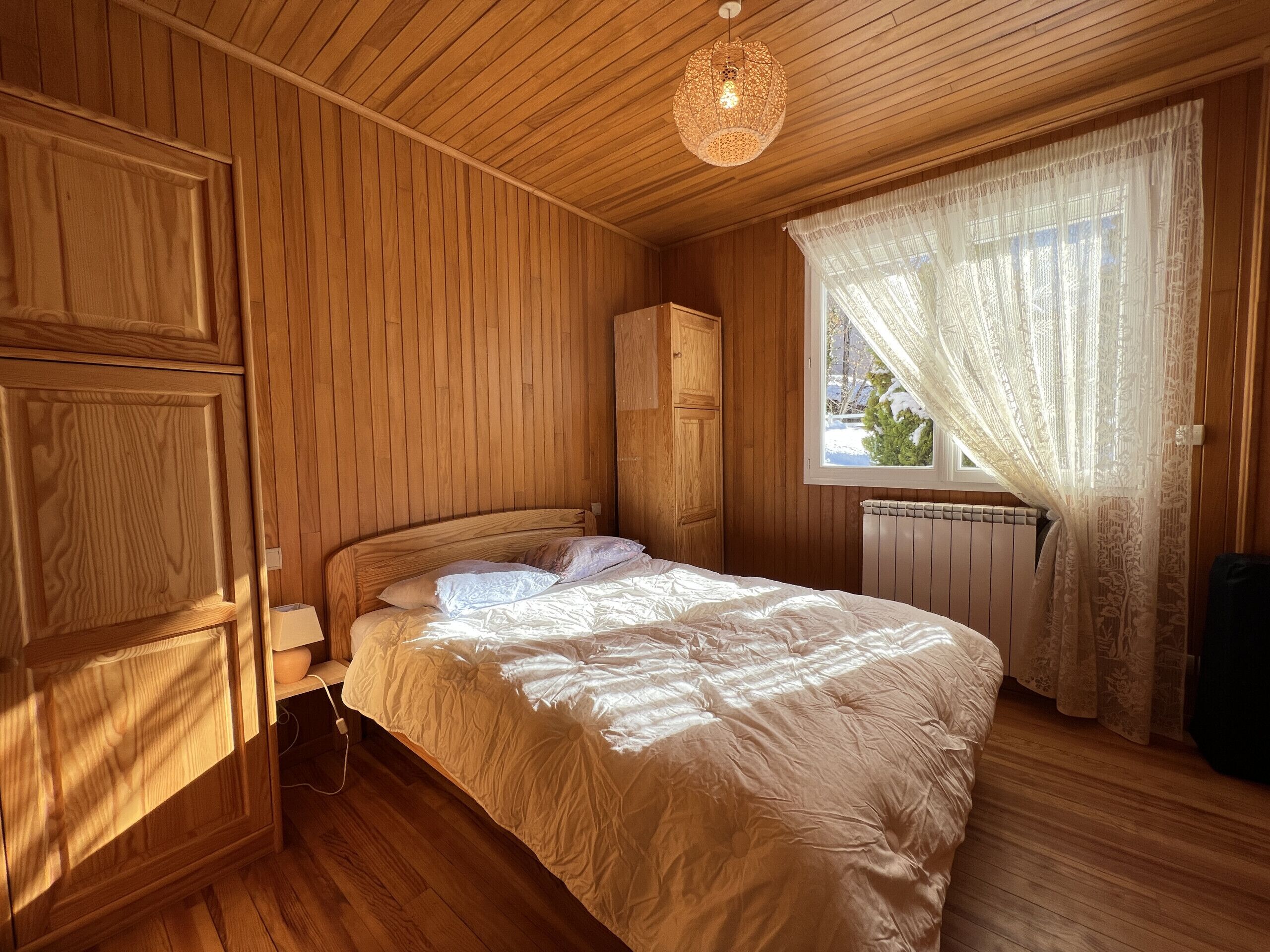 Een gezellige houten kamer met een bed, een radiator en een raam met dunne gordijnen.