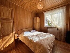 A cozy wooden room with a bed, a radiator, and a window with sheer curtains.