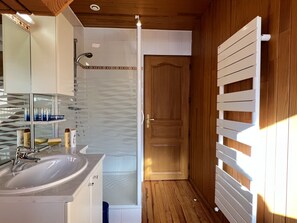 A bathroom with a wooden floor, a glass shower enclosure, a white sink, and a wooden door.