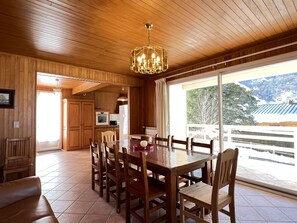 A wooden interior with a dining table and chairs, a kitchen area, and a large window with a view of a snowy landscape.