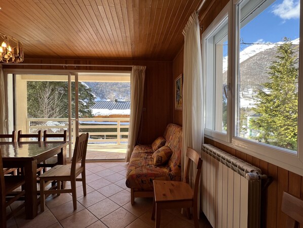 A cozy room with a wooden ceiling, a dining table with chairs, a sofa with patterned cushions, and a large window offering a view of snow-capped mountains and trees.