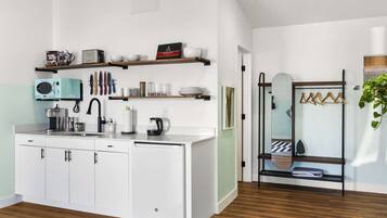 Una cocina moderna con armarios blancos, microondas integrado y cafetera. El piso es de madera y hay una puerta de vidrio que da a un baño.