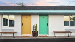 A building with two doors, one yellow and one green, each with a number above the door. There are two benches in front of the doors. - SCH04 · Mokulua Studio suite. hōʻoluʻolu (Chandler)