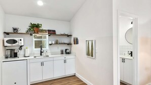 A modern kitchen with white cabinets, a microwave, coffee maker, and a sink. There are shelves with decorative items and a framed picture on the wall. - Studio Unit.hōʻoluʻolu (Chandler)