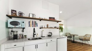 Una cocina moderna con armarios blancos, microondas, cafetera y hervidor. Hay estantes sobre la encimera con diversos objetos.