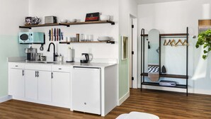 Una cocina moderna con armarios blancos, microondas integrado y cafetera. El piso es de madera y hay una puerta de vidrio que da a un baño.