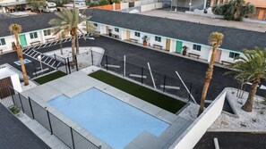 Un espace piscine doté d’une piscine rectangulaire, de palmiers et d’une rangée de bâtiments de style motel, avec un toit plat et des bordures bleu clair.