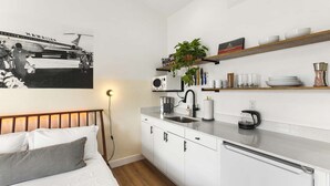 Une cuisine moderne avec des meubles blancs, une machine à café et un évier. On y trouve un lit avec une tête de lit et des oreillers, ainsi qu’une étagère murale où sont rangés des ustensiles de cuisine.