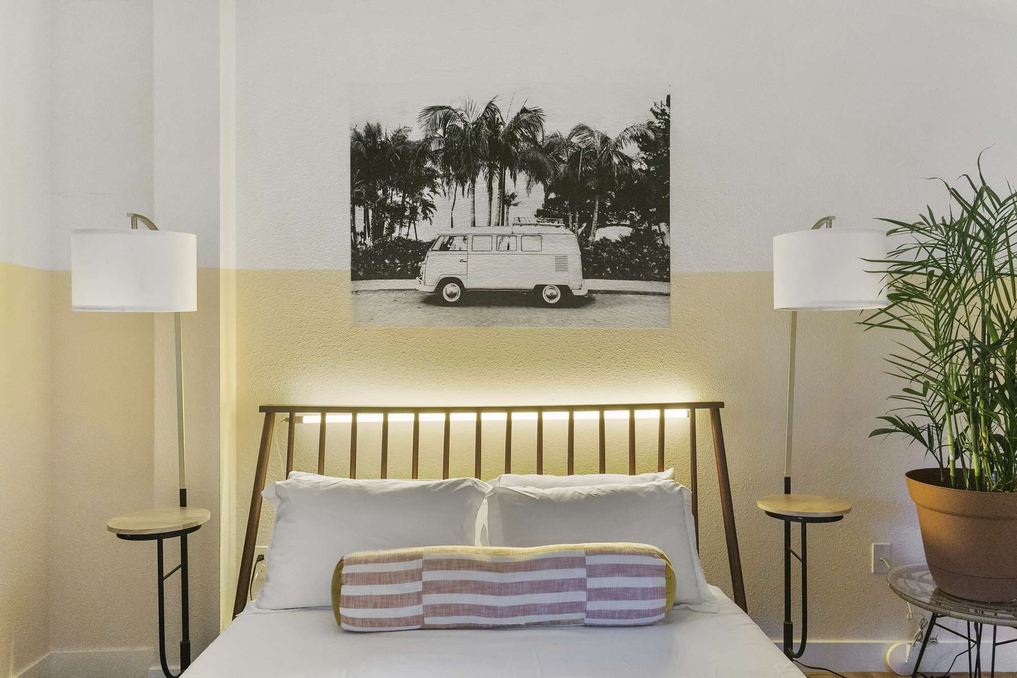Une chambre à coucher avec un lit, deux tables de chevet, une lampe, une plante en pot et un tableau représentant un camion, accroché au mur.