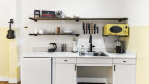 Een kleine keuken met witte kastjes, een zwarte gitaar, een koffiezetapparaat, een waterkoker en een magnetron.
