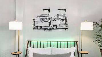 Un dormitorio con una cama, dos mesitas de noche, un cabecero, una lámpara de pared, una obra de arte con un camión y una planta en maceta.