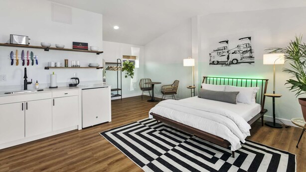 A modern hotel room with a bed, bedside table, lamp, and a kitchen area with cabinets and a sink. - SCH24 - Pohakulua Studio suite.nui (Chandler)