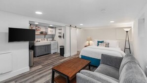 A modern hotel room with a kitchenette, a flat-screen TV, a bed with blue bedding, a grey sofa, and a wooden coffee table. - CC01 - Columbia City Urban Studio (Seattle)