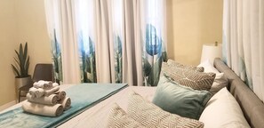 Una habitación de hotel con cama, almohadas, una toalla enrollada, una planta y cortinas con estampado de hojas.