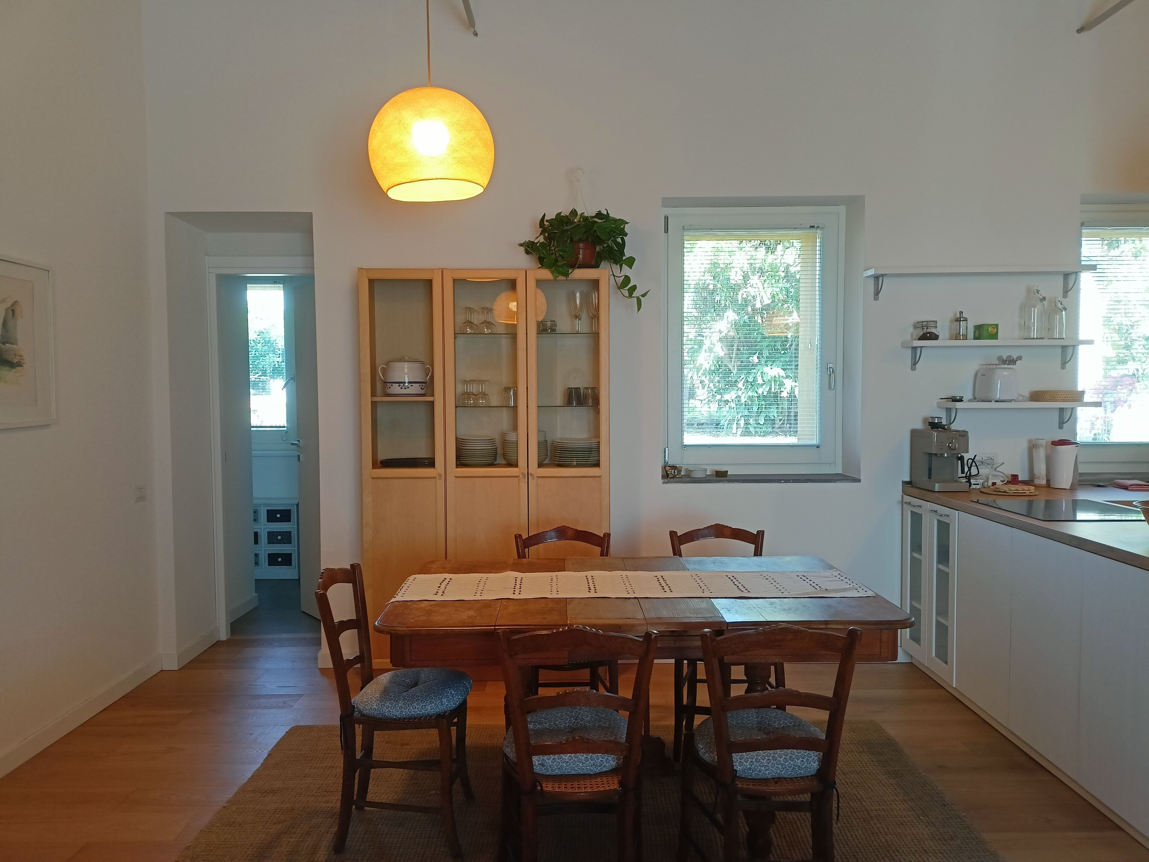 A dining area with a wooden table and chairs, a hanging light fixture, a window with blinds, and a kitchen area with shelves and appliances.