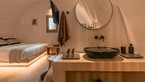 A bedroom with a bed, bedside table, mirror, and a bathroom with a sink and toiletries. - Living Moments (Santorini)