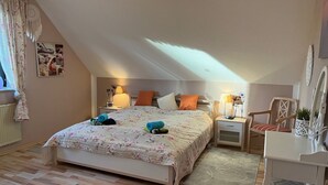 Et hyggeligt soveværelse med skrå vægge, en seng med blomstret sengetøj, to sengeborde med lamper, en stol og et lille skrivebord med et spejl.