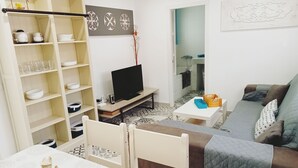 Ein Wohnzimmer mit Fernseher, Sofa und Esstisch. Ein Bücherregal mit diversen Gegenständen und einem Wanddekor.