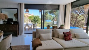 A modern living room with a sofa, a dining table, and chairs. There is a view of the outdoors through large windows.