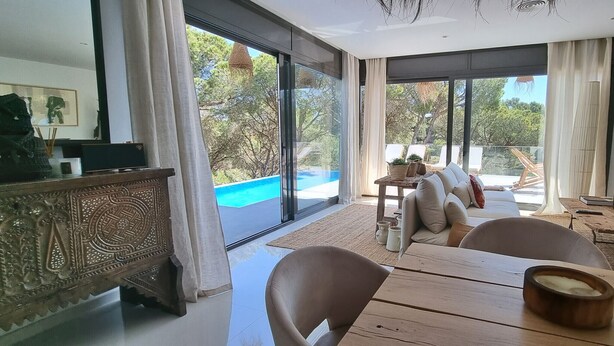 A modern living room with a large sliding glass door leading to a balcony, a dining area with a wooden table, and a flat-screen TV mounted on the wall.