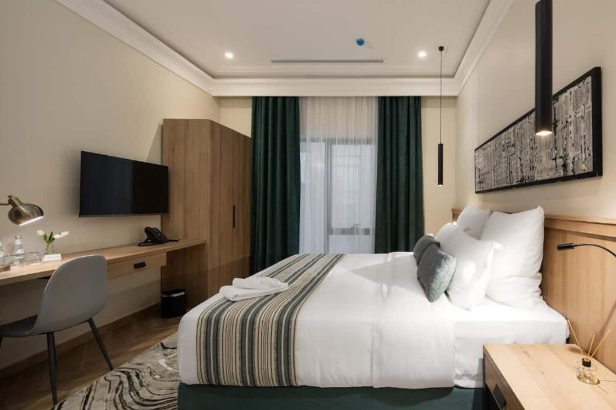 A hotel room with a bed, a desk with a chair, a television, and a large window with curtains.