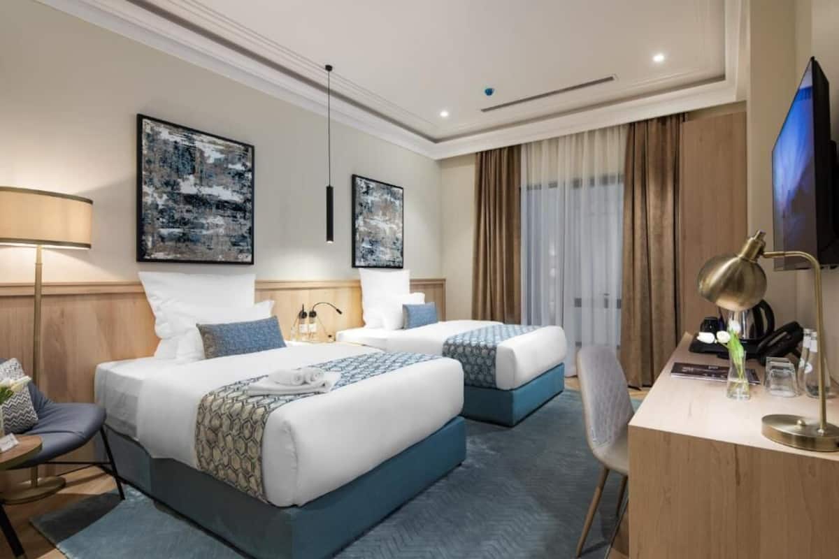 A hotel room with two beds, a desk, a TV, and artwork on the walls.