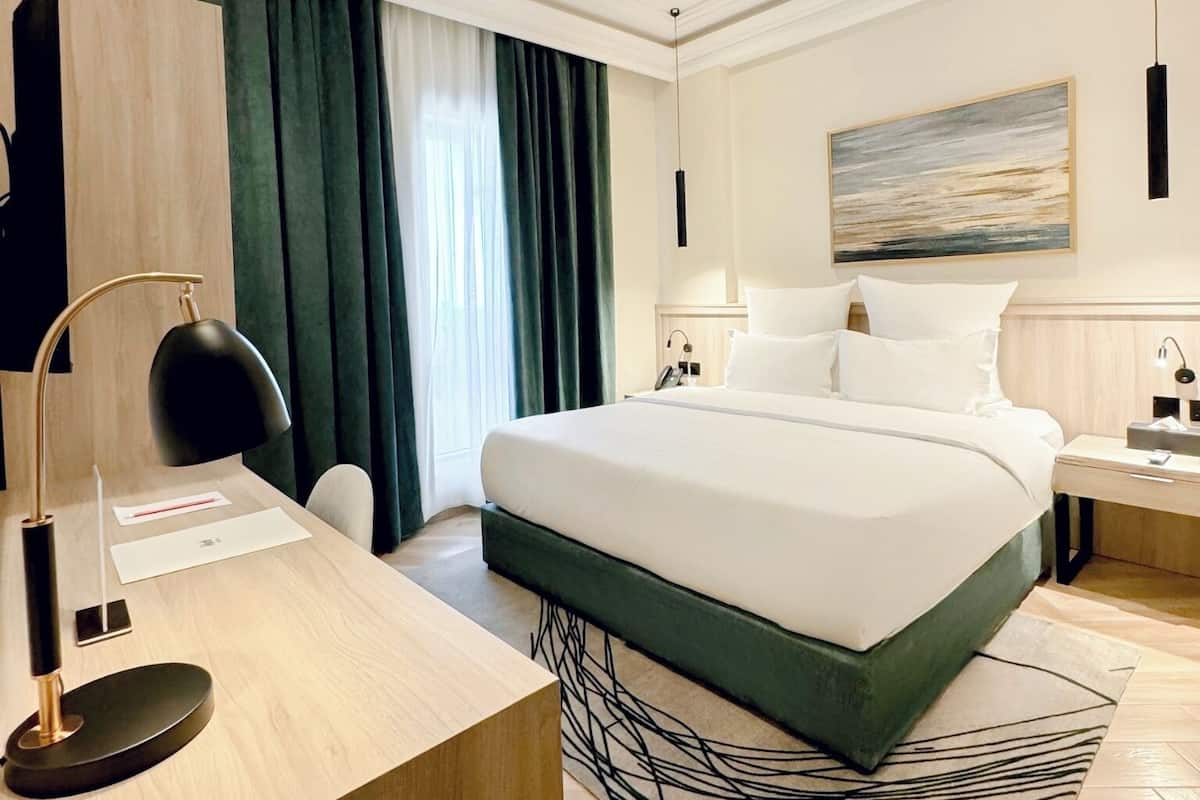 A modern hotel room with a large bed, a desk with a lamp, a bedside table, and a painting on the wall.