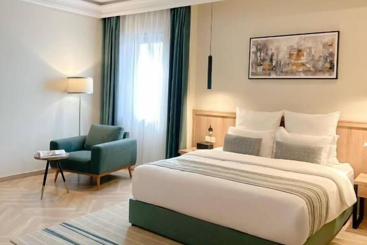 A modern hotel room with a large bed, a green armchair, a side table, a lamp, and a painting on the wall.